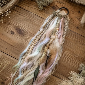 Könnte beinhalten: Ein Haaraccessoire mit einem schwarzen Gummiband, an dem eine Ansammlung geflochtener und gewellter Kunsthaarsträhnen in Pastelltönen wie Rosa, Blau und Creme befestigt ist, verziert mit Holzperlen, Federn und einem kleinen Libellen-Anhänger aus Metall.