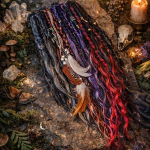 Op de afbeelding: Een verzameling kleurrijke dreadlocks met rode, paarse en zwarte lokken, versierd met veren en kralen. De dreadlocks zijn gerangschikt op een stenen oppervlak met een mystieke, bohemien uitstraling. Een brandende kaars en een schedel zijn op de achtergrond.