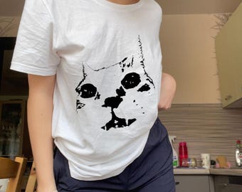 Könnte beinhalten: Weißes T-Shirt mit einer schwarzen Katzenkopf-Grafik. Die Augen und die Nase der Katze sind schwarz, mit einer einfachen Umrisslinie des Katzenkopfes. Das Shirt ist kurzärmlig und wahrscheinlich aus Baumwolle.
