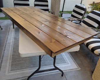 May include: A rectangular wooden table with a light brown finish, resting on a white folding table with black legs. The table is surrounded by black and white striped chairs, suggesting outdoor dining or event setup.