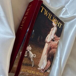 Peut inclure: Un livre relié intitulé Twilight de Stephenie Meyer avec une tranche en velours rouge foncé et un marque-page ruban. La couverture présente une peinture classique d'une personne debout près d'un lion et d'un agneau, avec des lettres dorées.
