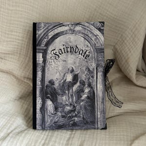 Op de afbeelding: Een rechthoekig hardcover boek met een zwarte rug en een zwart kanten lint. De voorkant toont een grijstinten illustratie van een klassieke architecturale boog die een religieuze scène met figuren en de tekst Fairndale omlijst.