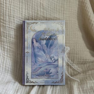 May include: A hardcover book titled When the Moon Hatched by Sarah A. Parker, featuring a blue and purple sky-themed cover with a central illustration of a figure, gold foil accents, a purple spine, and a white lace ribbon tied to the side.