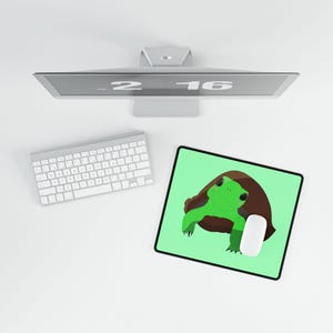 Puede incluir: Un equipo informático con un monitor plateado que muestra la hora 2:16, un teclado inalámbrico blanco y una alfombrilla de ratón rectangular con fondo verde claro, una ilustración de una tortuga verde con caparazón marrón y un ratón blanco.