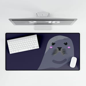 May include: A rectangular desk mat featuring a dark blue background and a large, stylised grey seal illustration. The seal has black eyes, a black nose, textured whiskers, and small pink circular cheeks. The mat is positioned under a white keyboard.
