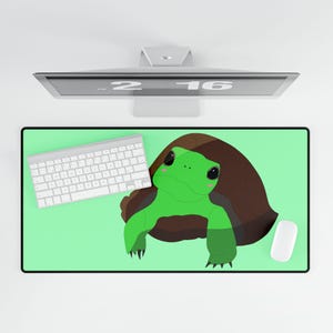May include: A rectangular desk mat featuring a light green background and a central illustration of a green turtle with a dark brown shell. The mat is positioned under a white computer keyboard and a white computer mouse, with a monitor visible above.