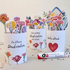 Puede incluir: Tres recipientes cilíndricos blancos decorados con ilustraciones florales y texto en alemán. Cada recipiente contiene recortes de flores de papel. Uno dice "Ein kleines Dankeschön", otro "Ein süßes Dankeschön für Dich" y el tercero "Vielen Dank". También hay un tubo de pegamento.