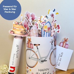 Peut inclure: Une carte d'anniversaire blanche avec une illustration de v&eacute;lo et les mots "Happy Birthday". La carte pr&eacute;sente un design pop-up avec un g&acirc;teau, des fleurs et une girafe. Le texte "bist richtig wertvoll" est visible sur un tube blanc.