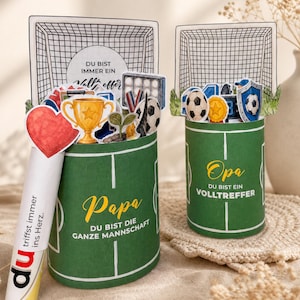 Könnte beinhalten: Zwei Geschenkbehälter zum Thema Fußball. Ein grüner Behälter sagt "Papa" und "DU BIST DIE GANZE MANNSCHAFT". Der andere sagt "Opa" und "DU BIST EIN VOLLTREFFER". Jeder hat ein Fußballtor und verschiedene Dekorationen.