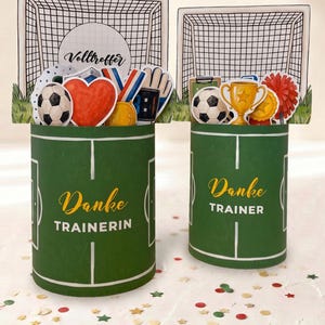Op de afbeelding: Twee groene cadeau dozen met voetbalthema met de woorden "Danke TRAINERIN" en "Danke TRAINER" in gouden letters. Elke doos heeft een voetbaldoel achtergrond en diverse voetbal gerelateerde decoraties.