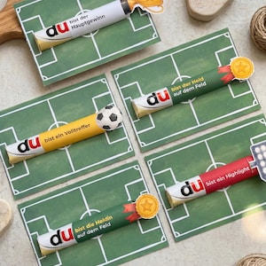 Könnte beinhalten: Fünf rechteckige Karten mit einer grünen Fußballfeld-Grafik, jede mit einer zylindrischen Süßigkeitenverpackung versehen. Die Verpackungen zeigen den Text 'du bist der Hauptgewinn', 'du bist der Held auf dem Feld', 'du bist ein Volltreffer', 'du bist ein Highlight' und 'du bist die Heldin auf dem Feld' mit angebrachten Papier-Symbolen eines Pokals, eines Fußballs, Sternmedaillen und Flutlichtmasten.
