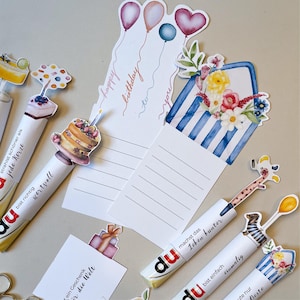 Peut inclure: Un assortiment de stylos d&eacute;coratifs et de cartes d'anniversaire. Les stylos pr&eacute;sentent divers motifs, dont un g&acirc;teau, une girafe et des fleurs. Les cartes comportent des illustrations de ballons et de c&oelig;urs, avec l'inscription "happy birthday to you".