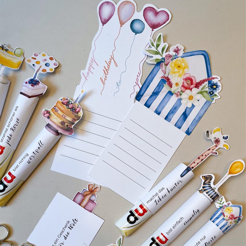 Peut inclure: Un assortiment de stylos d&eacute;coratifs et de cartes d'anniversaire. Les stylos pr&eacute;sentent divers motifs, dont un g&acirc;teau, une girafe et des fleurs. Les cartes comportent des illustrations de ballons et de c&oelig;urs, avec l'inscription "happy birthday to you".