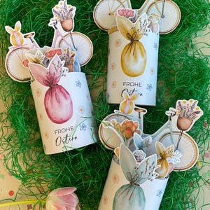 Op de afbeelding: Paasdecoraties met cilindrische containers versierd met bloemen- en konijnenillustraties, vergezeld van fietscut-outs met bloemenmanden. De containers hebben de tekst "FROHE Ostern" en staan op groen decoratief gras.