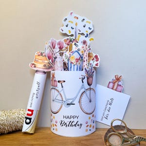 Peut inclure: Un pr&eacute;sentoir &agrave; cartes d'anniversaire blanc avec une illustration de v&eacute;lo et les mots "Happy Birthday". Le pr&eacute;sentoir est d&eacute;cor&eacute; de confettis color&eacute;s et surmont&eacute; de d&eacute;coupes de fleurs, d'un g&acirc;teau et de cadeaux. Un stylo avec du texte est &agrave; gauche.