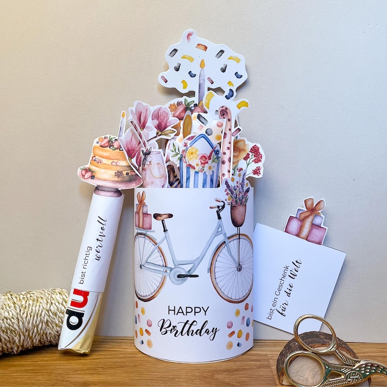 Peut inclure: Un pr&eacute;sentoir &agrave; cartes d'anniversaire blanc avec une illustration de v&eacute;lo et les mots "Happy Birthday". Le pr&eacute;sentoir est d&eacute;cor&eacute; de confettis color&eacute;s et surmont&eacute; de d&eacute;coupes de fleurs, d'un g&acirc;teau et de cadeaux. Un stylo avec du texte est &agrave; gauche.