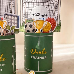 Op de afbeelding: Groene cilindrische geschenkdoos met een voetbalveldontwerp, met de tekst "Danke TRAINER". De doos is versierd met voetbalthema-uitsparingen, waaronder een voetbal, een trofee en een doelnet. Het woord "Volltreffer" is ook zichtbaar.
