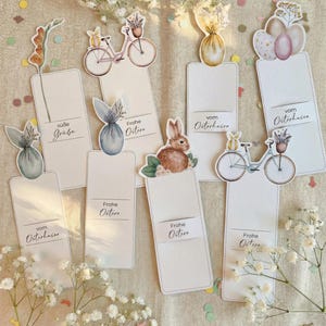 May include: A collection of eight rectangular paper gift tags featuring watercolor illustrations of Easter themes including bicycles, rabbits, and eggs. The tags display German text such as "Frohe Ostern," "süße Grüße," and "vom Osterhasen" printed on white cardstock.
