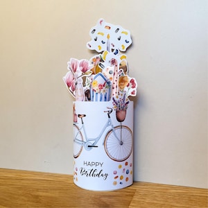 Peut inclure: Carte d'anniversaire pop-up cylindrique blanche avec une illustration de v&eacute;lo bleu clair et un arrangement floral. La carte pr&eacute;sente une girafe et les mots "Happy Birthday". La base est d&eacute;cor&eacute;e de points de confettis color&eacute;s.