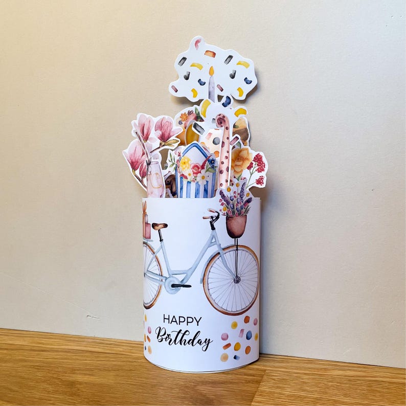Peut inclure: Carte d'anniversaire pop-up cylindrique blanche avec une illustration de v&eacute;lo bleu clair et un arrangement floral. La carte pr&eacute;sente une girafe et les mots "Happy Birthday". La base est d&eacute;cor&eacute;e de points de confettis color&eacute;s.