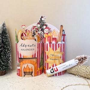 Könnte beinhalten: Ein zylindrischer Adventskalender mit rot-weißen Streifen, Kamin-Illustration und Weihnachtsmotiven, beschriftet mit Advents KALENDER, 24 kleine Freuden sowie du bist einfach wunderbar auf einer kleinen Röhre mit Tannenzapfen-Grafik.