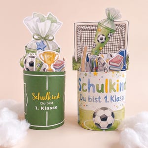 May include: Two cylindrical containers featuring football-themed decorations. The left one is green with white lines and text reading Schulkind Du bist 1. Klasse. The right one is white with confetti, a football, and text reading Schulkind Du bist 1. Klasse.