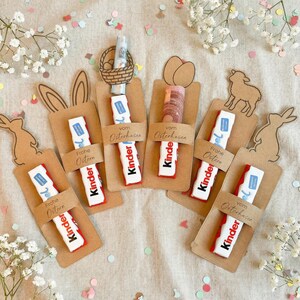 May include: An assortment of Easter-themed gifts, including Kinder chocolate bars, arranged on a beige surface. Each gift is attached to a brown card with decorative cutouts of Easter bunnies, eggs, and a lamb. The words "Frohe Ostern" and "vom Osterhasen" are visible.
