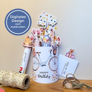 Peut inclure: Un cylindre blanc avec une illustration de v&eacute;lo et l'inscription "Happy Birthday". Le cylindre est orn&eacute; de d&eacute;coupes de papier de fleurs, d'un g&acirc;teau et d'un cadeau. Un stylo et une carte avec du texte sont &eacute;galement pr&eacute;sents.