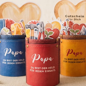 May include: Three cylindrical containers in blue, red, and yellow, each with the word "Papa" in white script. The containers are filled with various stickers, including tools, hearts, and a sun. The text "DU BIST DER HELD FÜR JEDEN EINSATZ" is printed on each container.