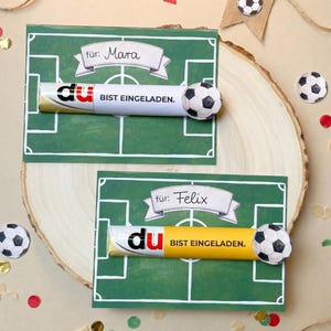 May include: Two green cards with a soccer field design, each with a candy bar and a soccer ball attached. One card says "f&uuml;r: Mara" and the other says "f&uuml;r: Felix". The candy bars have the text "du BIST EINGELADEN."