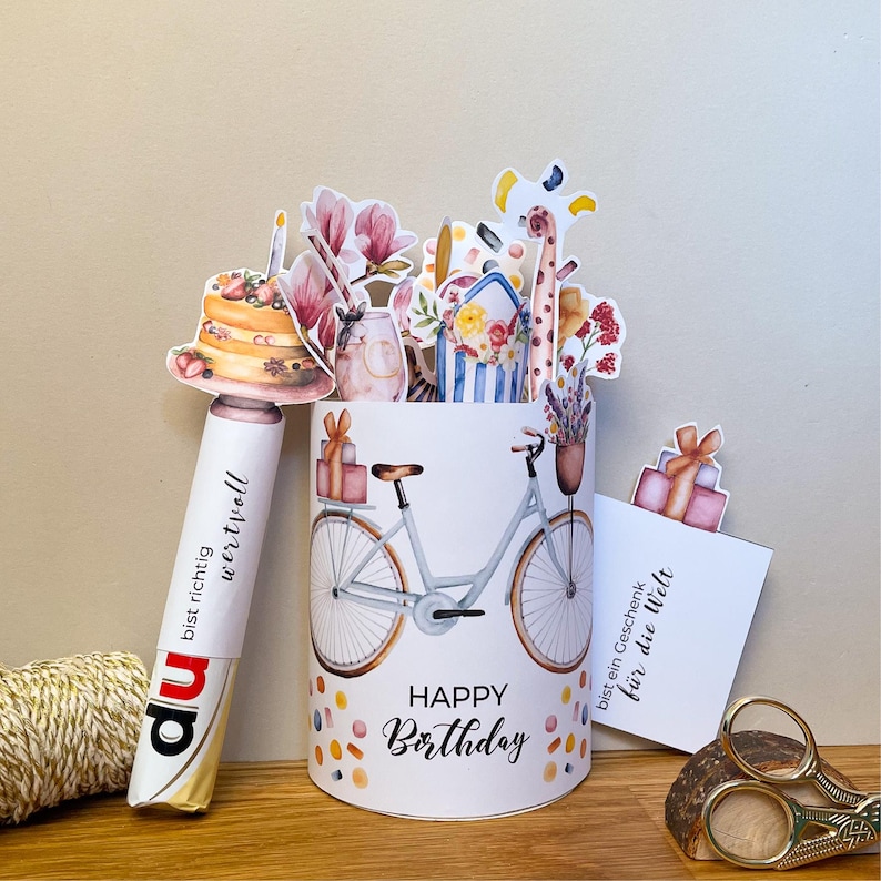 Könnte beinhalten: Ein weißer Geburtstagskartenhalter mit einer Fahrradillustration und dem Schriftzug "Happy Birthday". Der Halter ist mit Konfetti und Pop-up-Blumen- und Geschenkbox-Dekorationen verziert. Ein Stift mit Text befindet sich links.