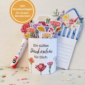 以下が含まれることがあります： 水彩画の花のイラストと「Ein s&uuml;&szlig;es Dankesch&ouml;n f&uuml;r Dich」というテキストが描かれた白い紙筒。 Duploキャンディーの棒と青と白のストライプの封筒も表示されています。画像には、カラフルな紙の花の切り抜きが含まれています。