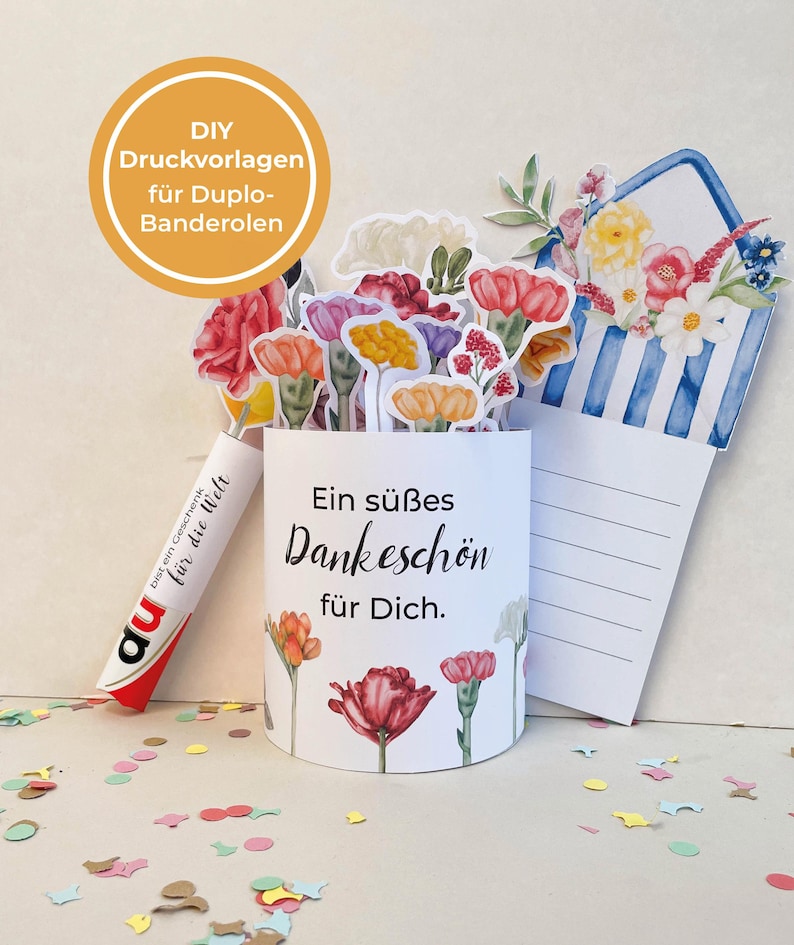 以下が含まれることがあります： 水彩画の花のイラストと「Ein s&uuml;&szlig;es Dankesch&ouml;n f&uuml;r Dich」というテキストが描かれた白い紙筒。 Duploキャンディーの棒と青と白のストライプの封筒も表示されています。画像には、カラフルな紙の花の切り抜きが含まれています。