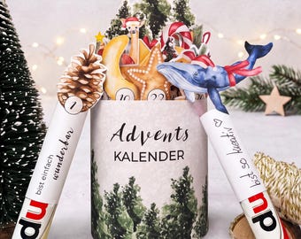 Op de afbeelding: Een cilindervormige adventskalender met een bosprint en de tekst Advents KALENDER. Het bevat pop-up illustraties van een dennenappel, een hert, een ster en een blauwe vinvis. Twee witte kokers zijn bevestigd, gelabeld du bist einfach wunderbar en du bist so kreativ.