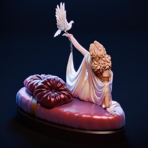 Puede incluir: Escultura de una mujer reclinada en una chaise longue roja, sosteniendo una paloma blanca. La mujer lleva un vestido blanco fluido y tiene el pelo rubio rizado. Dos cojines rojos y redondos est&aacute;n en la chaise longue. La escultura est&aacute; sobre un fondo oscuro.