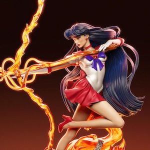 May include: A detailed figurine of a character in a red and white sailor uniform with a blue bow. The figure has long, dark blue hair and is posed with a fiery weapon. The base of the figure has Japanese text.
