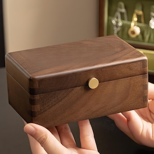 May include: A rectangular wooden jewelry box with a dark brown finish and a small, round, gold-colored knob. The box is held by two hands, and the wood grain is visible. The box is likely for storing jewelry.