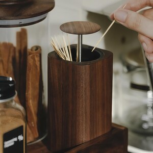 May include: A dark brown wooden toothpick holder with a matching lid. The holder is square with rounded edges and contains several toothpicks. A hand is reaching in to grab a toothpick. The holder sits on a wooden shelf.