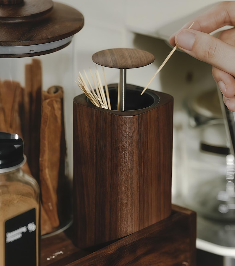 May include: A dark brown wooden toothpick holder with a matching lid. The holder is square with rounded edges and contains several toothpicks. A hand is reaching in to grab a toothpick. The holder sits on a wooden shelf.