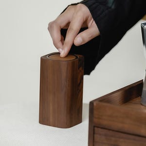 May include: A dark brown wooden container with a square top and rounded corners. The lid is being opened by a person's finger. The container is sitting on a white surface. The container is part of a set of wooden items.