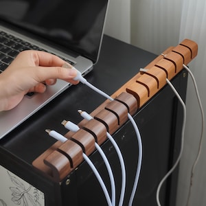 Op de afbeelding: Een houten kabelorganizer met meerdere sleuven, met witte oplaadkabels. De organizer is gemaakt van twee tinten hout, met een laptop op de achtergrond. Een hand houdt een witte kabel vast.
