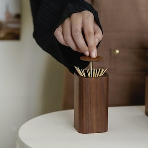 May include: A wooden toothpick holder with a dark brown finish. The holder is rectangular with rounded corners and a small, round lid. The lid is being lifted to reveal the toothpicks inside. The holder sits on a white surface.