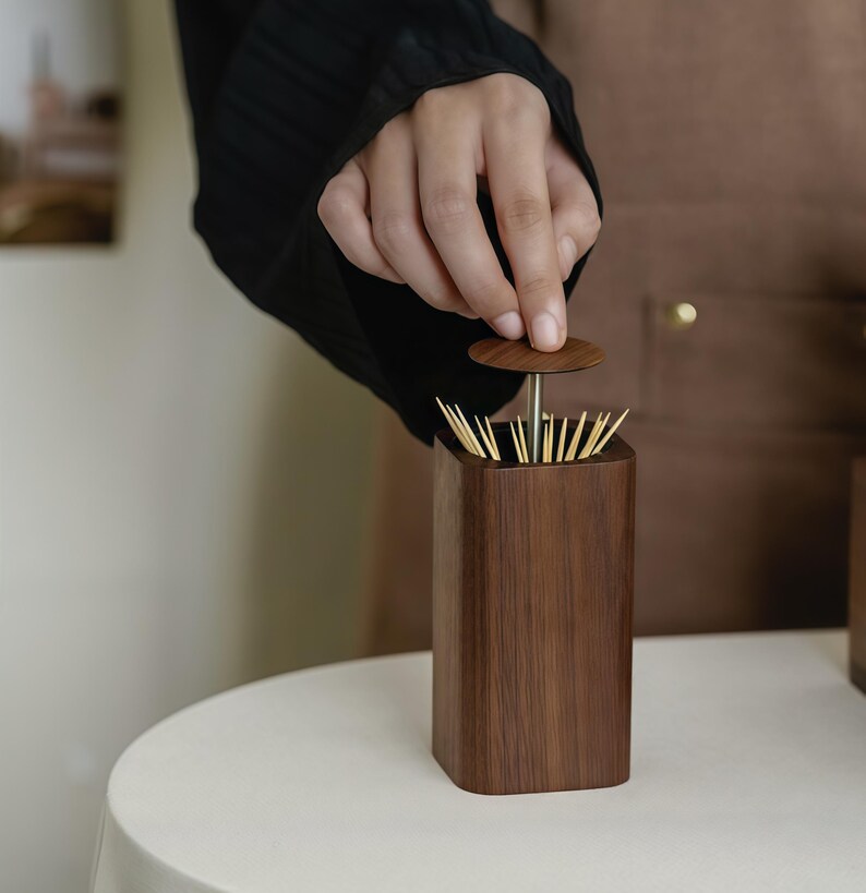 May include: A wooden toothpick holder with a dark brown finish. The holder is rectangular with rounded corners and a small, round lid. The lid is being lifted to reveal the toothpicks inside. The holder sits on a white surface.