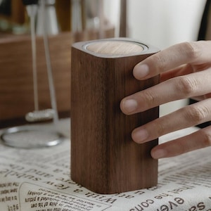 May include: A dark brown wooden container with a light brown wooden top. The container has a rectangular shape with rounded corners. The container is being held by a person's hand.