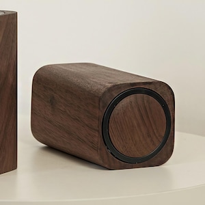 May include: A rectangular wooden speaker with rounded corners and a dark brown finish. The speaker has a circular black accent on one end. The wood grain is visible. The speaker is on a white surface.