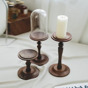 May include: Three dark brown wooden candle holders. One has a clear glass cloche, another holds a white candle, and the third is empty. The holders have a round base and a turned stem, creating a vintage aesthetic.