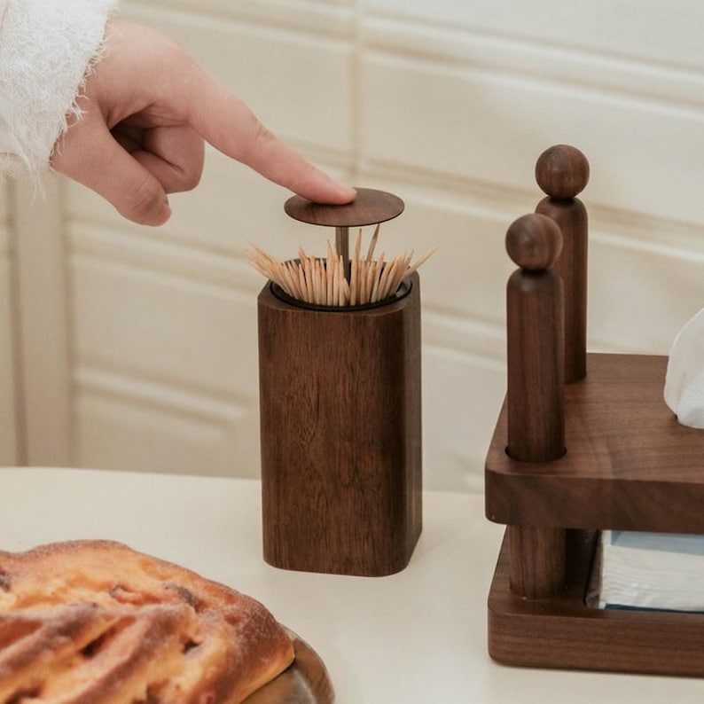 May include: A dark brown wooden toothpick dispenser with a circular lid. The dispenser is rectangular with rounded corners and contains numerous toothpicks. The lid is being pressed down by a finger. A wooden stand with two decorative figures is in the background.