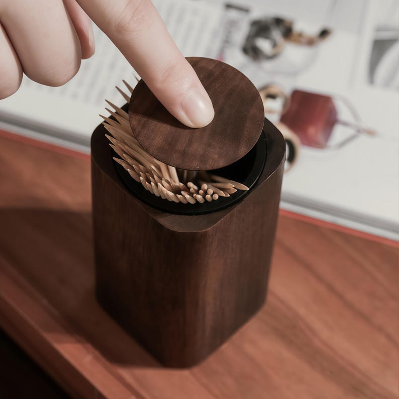 May include: A dark brown wooden toothpick holder with a round lid. The lid is being lifted to reveal the toothpicks inside. The holder has a square base with rounded corners and a black interior.