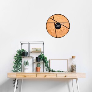 May include: A wooden desk with three drawers and white legs. A black metal shelf with plants and a white frame with a blank white background are on the desk. A black and wood clock with a geometric design is on the wall.