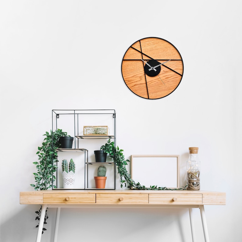 May include: A wooden desk with three drawers and white legs. A black metal shelf with plants and a white frame with a blank white background are on the desk. A black and wood clock with a geometric design is on the wall.
